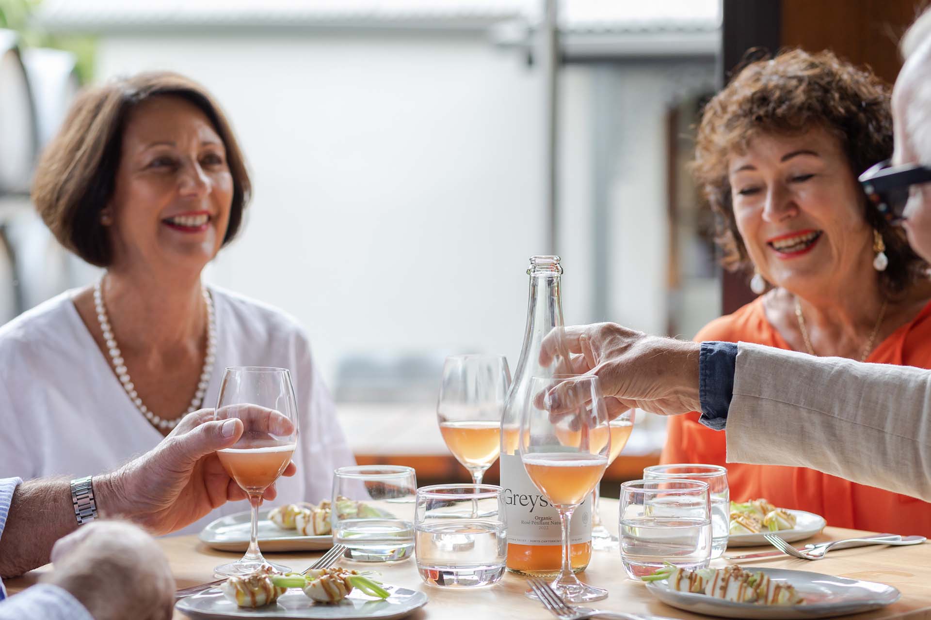 A photograph of two elderly couples sitting at a well-lit table. They are smiling while one of the men reachers for a bottle of Greystone wine placed at the center of the table. Each person has a glass of wine sat in front of them. 