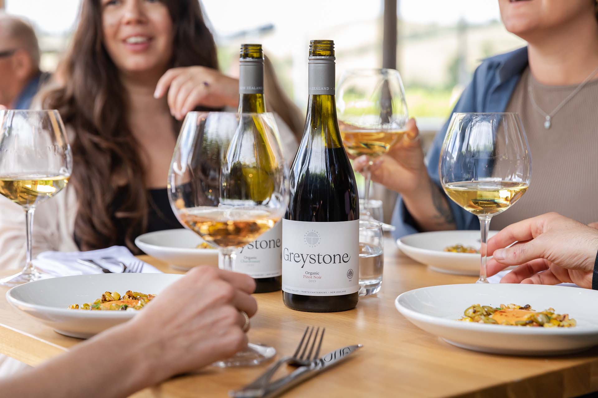 A group of three people are photographed enjoying a meal. Two bottles of Greystone wine are placed at the center of the table. Each person photographed has a glass of white wine sitting in front of them. 