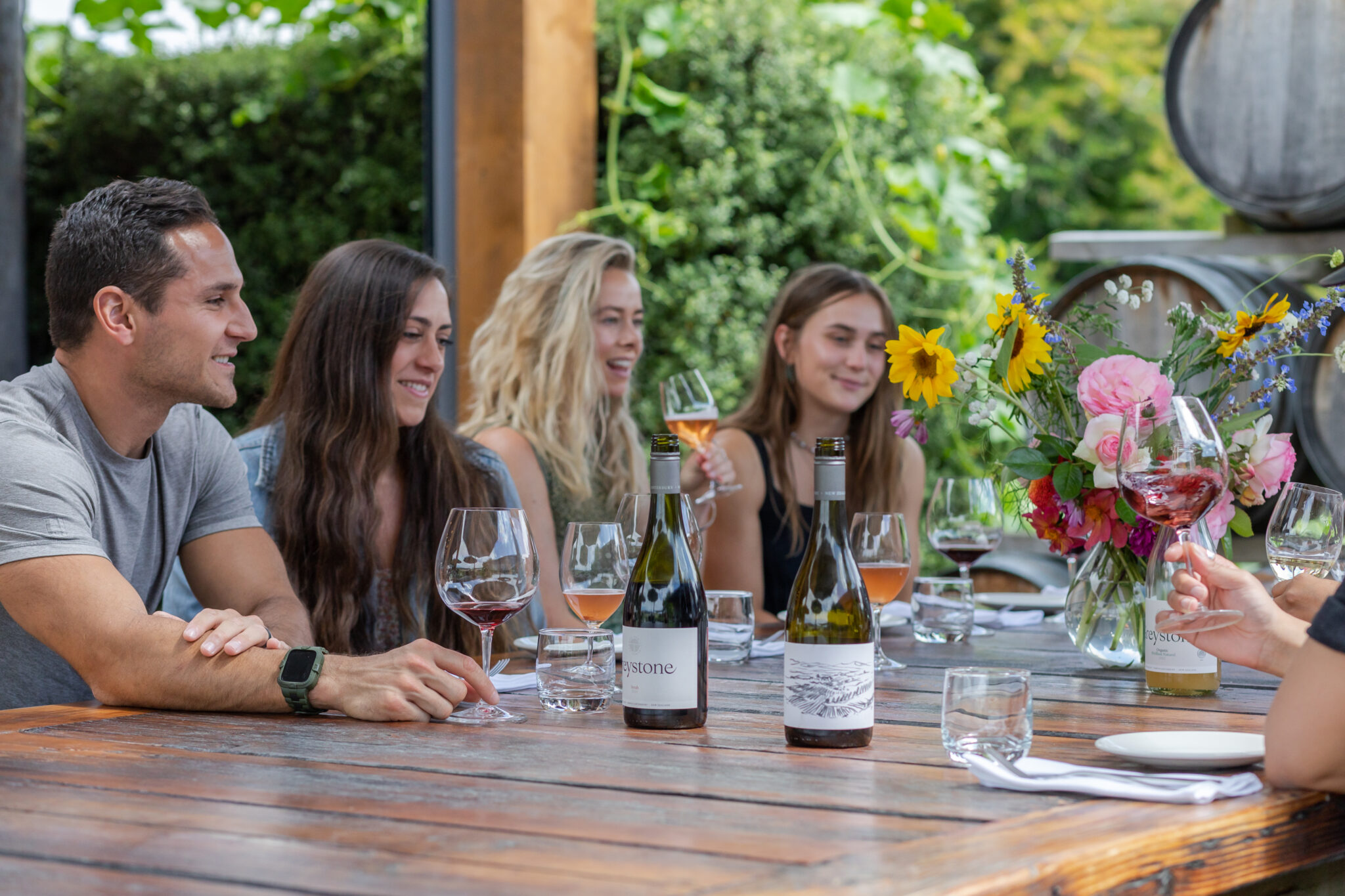 Group of friends enjoying wine tasting at Greystone Winery in North Canterbury, New Zealand. Outdoor dining with Greystone Pinot Noir and Chardonnay, surrounded by vineyards and wine barrels. Scenic winery experience with local, organic wines